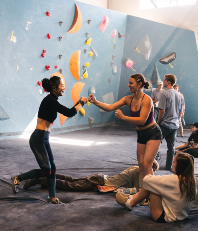 Two women in athletic wear smile and fist-bump in an indoor rock climbing gym, surrounded by others. Colorful climbing holds brighten the walls—perfect for anyone exploring a beginner’s guide to bouldering.