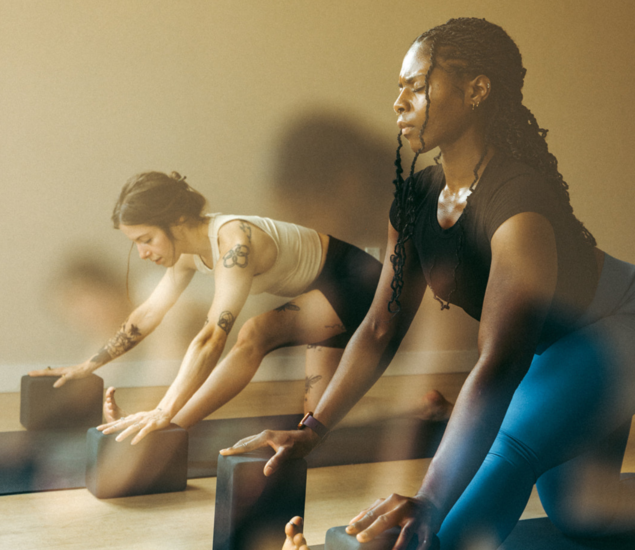 Two women in athletic wear practice yoga indoors, using yoga blocks to support a forward bend pose. Warm light softly illuminates the calm and focused atmosphere of yoga classes Brooklyn locals love.