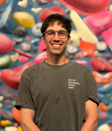 A smiling person wearing glasses and a Seattle Bouldering Project Staff t-shirt stands in front of a colorful indoor climbing wall, ready to help new climbers make the most of their summer membership.