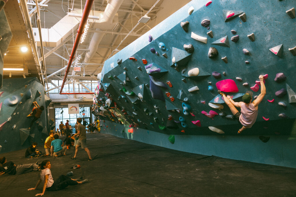 People enjoy bouldering and watching others at an indoor climbing gym, with colorful holds on angled walls and natural light streaming from above. Some climbers are actively scaling the walls, getting a full body workout, while others sit or stand nearby.