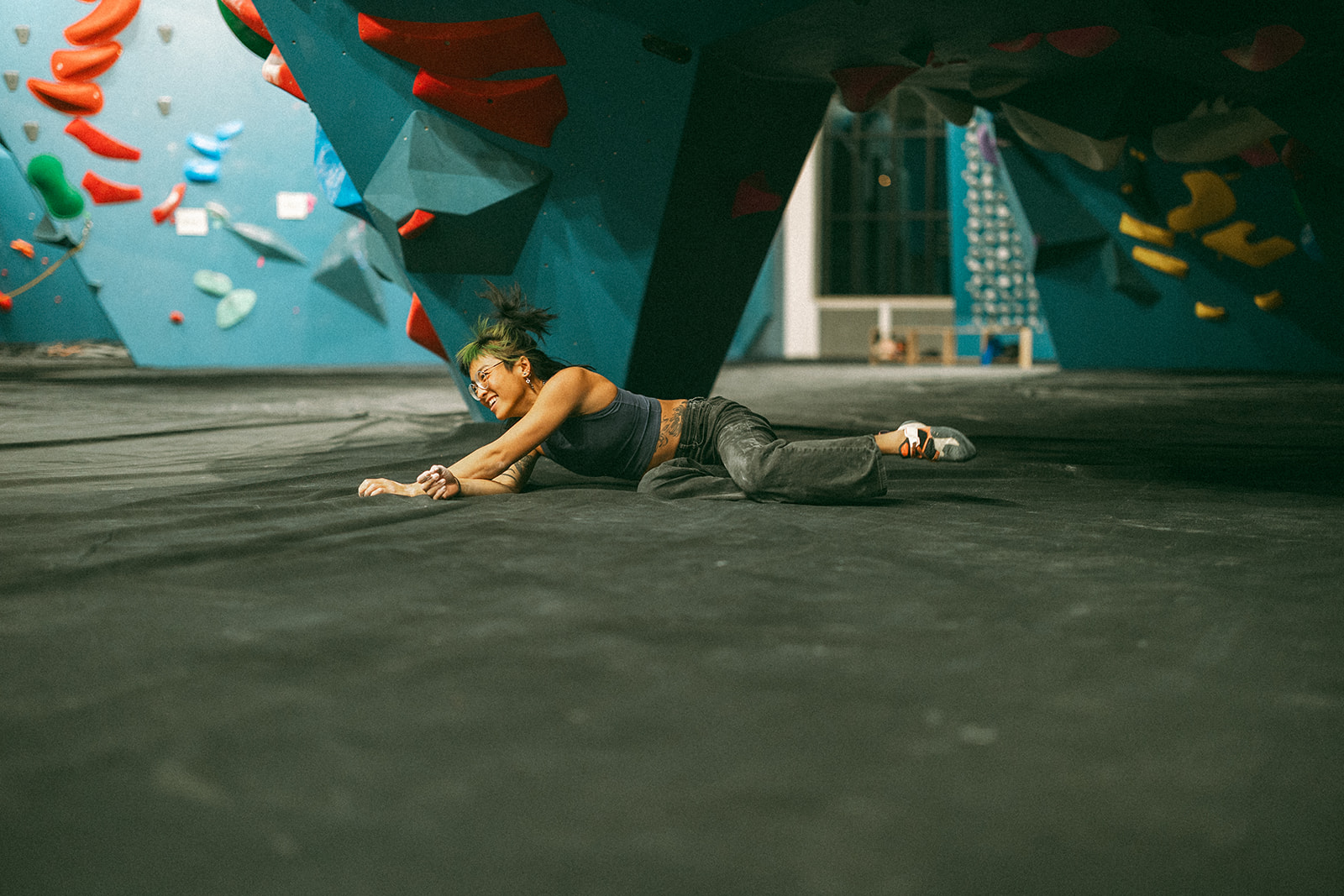 A person lies on the padded floor of an indoor rock climbing gym, reaching forward with one arm extended, surrounded by colorful holds that cover vibrant blue walls.