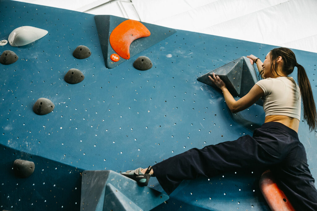 A woman wearing a beige top and dark pants climbs an indoor bouldering wall, gripping large blue and gray holds while stretching her leg to balance—a dynamic full body workout among the best workouts for strength and agility.