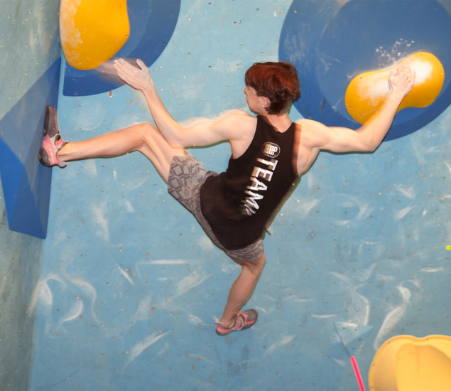 A person wearing a black TEAM tank top and gray shorts is climbing an indoor bouldering wall at the National Skills Camp, stretching between large yellow and blue holds in a Twin Cities simulation setting.