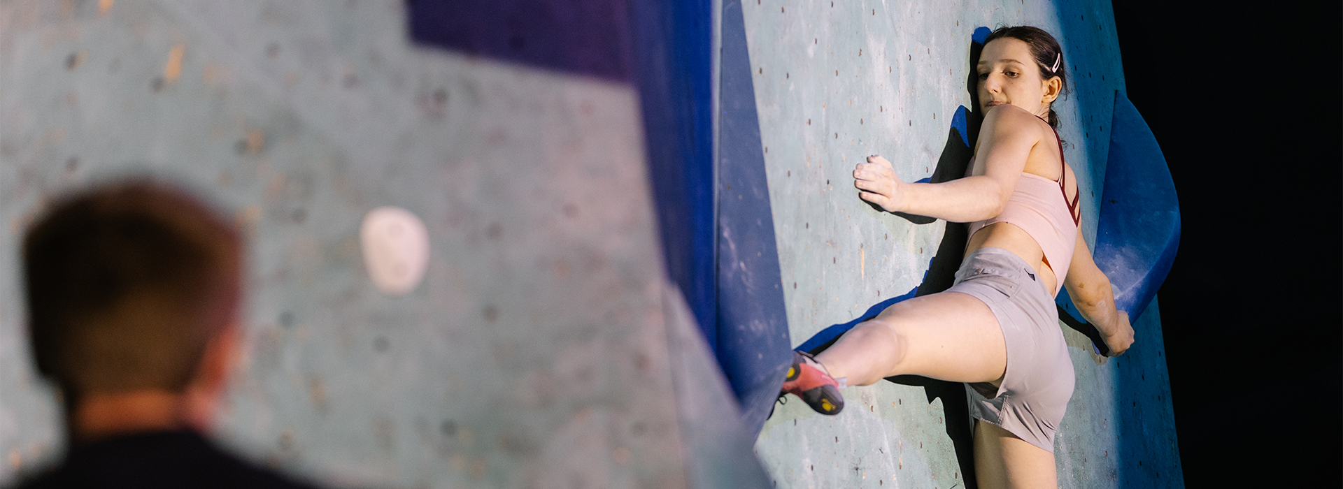 A woman in athletic clothing climbs an indoor bouldering wall, stretching her left leg wide for balance at a National Skills Camp. She looks focused, chalk on her hands, while a blurred figure watches in the foreground.