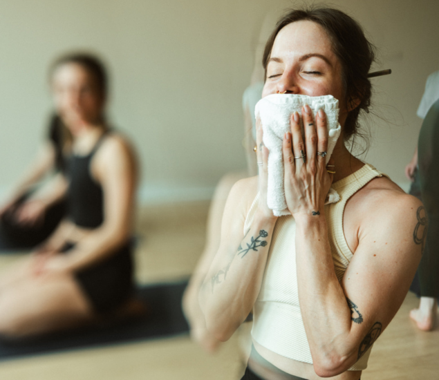 A woman in athletic wear holds a towel to her face with her eyes closed, appearing relaxed. Another person sits cross-legged on a yoga mat in the background, capturing the peaceful atmosphere of yoga classes Brooklyn offers.