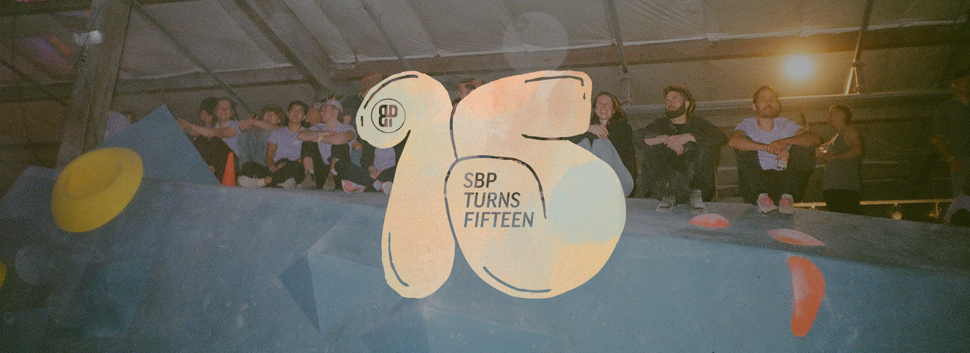 A group of people sit atop an indoor climbing wall during a Seattle climbing competition. Overlaid text reads “SBP Turns Fifteen” with a stylized “15.” The lighting is dim and festive, adding to the celebration.