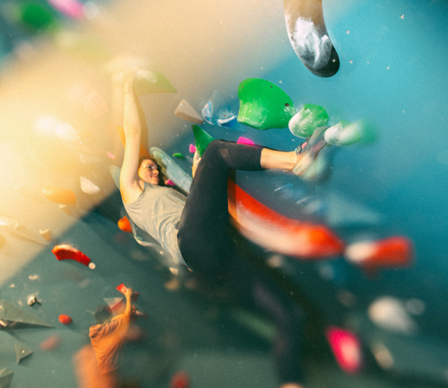 A person climbs an indoor bouldering wall at a Boston climbing gym, gripping colorful holds. The image is dynamic, with motion blur and warm light effects, capturing energy and the excitement of a summer membership.