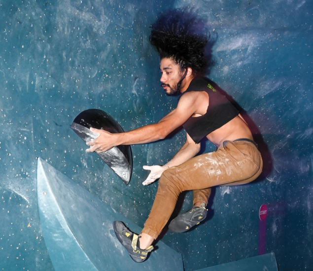 A person with long hair and a beard, wearing a black crop top and brown pants, climbs an indoor bouldering wall in Austin, gripping a black hold and appearing airborne with chalk on their hands.