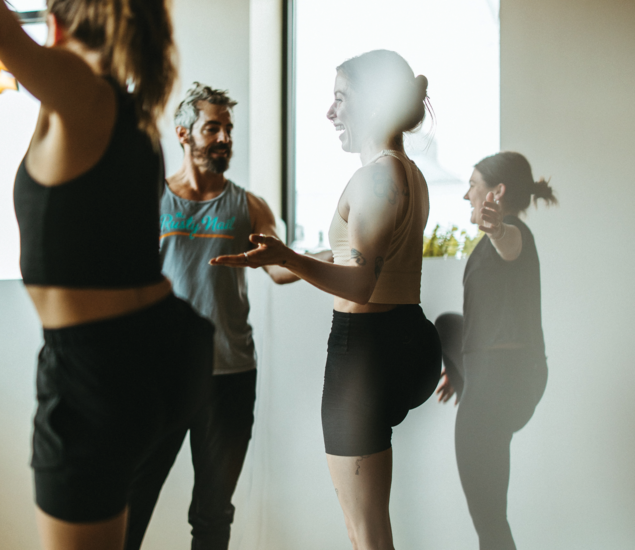 A group of people in athletic wear smile and talk together in a sunlit room, possibly during yoga classes Austin offers, with large windows and relaxed, friendly body language.
