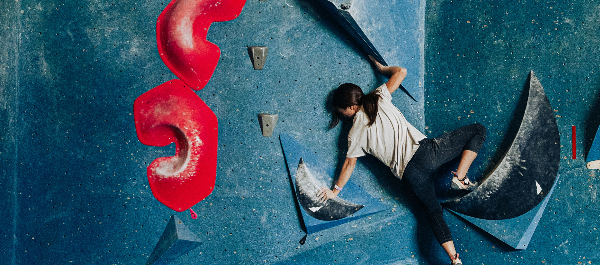 A person in a white shirt and dark pants is indoor rock climbing on a blue wall with large red and black handholds at an indoor climbing Seattle gym, reaching up with one arm while gripping a blue triangular hold with the other.