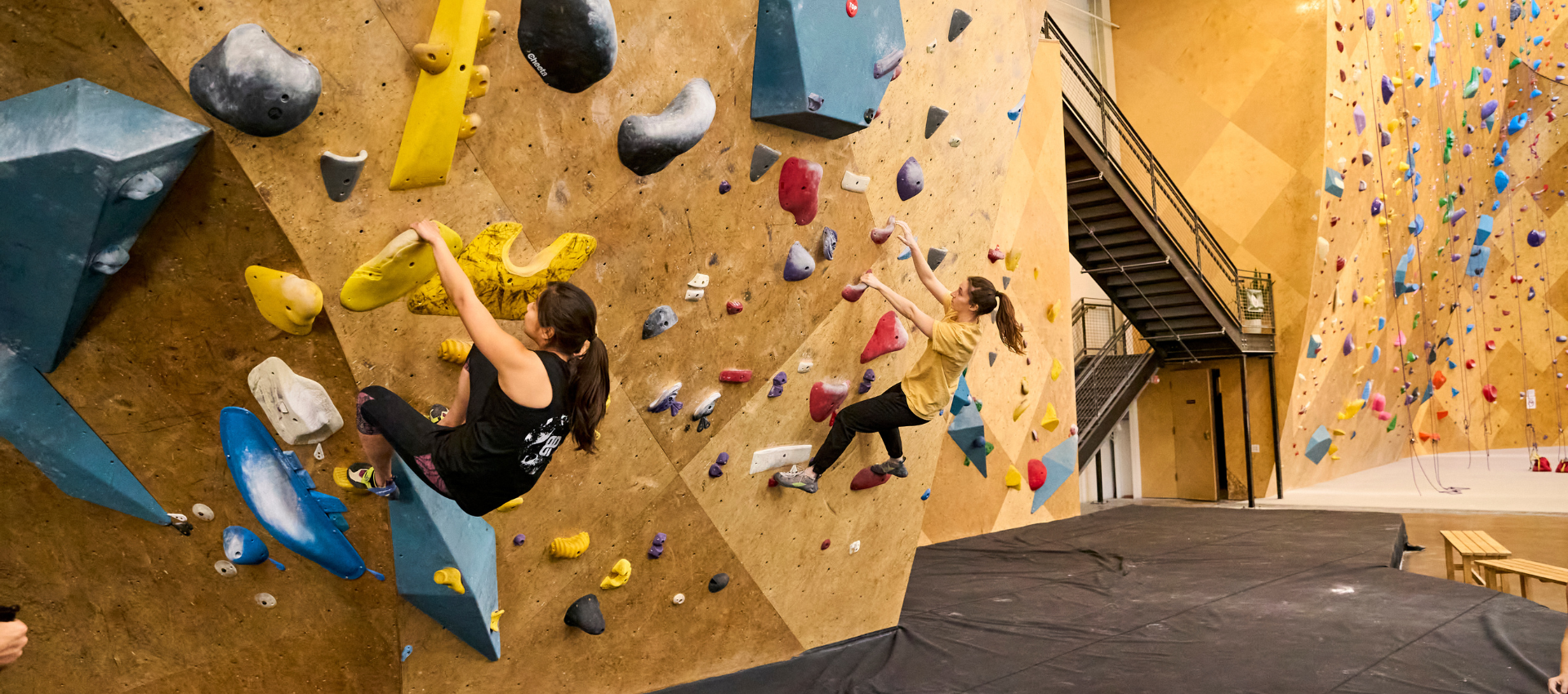 Two people are bouldering on an indoor climbing wall with colorful holds, climbing shoes, and safety mats below. The scene highlights climbing differences from rope climbing, with a staircase and more routes in the background.