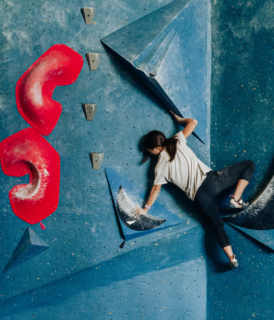A woman in athletic clothing is bouldering on an indoor climbing wall in Seattle, gripping blue holds with both hands and feet while two large red holds are visible to her left.