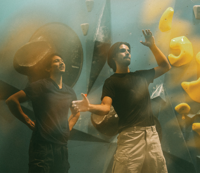Two people stand in front of an indoor climbing wall with yellow holds. One gestures with his hands, appearing to explain something about climbing classes in Austin, while the other smiles and listens. Both are dressed casually in dark shirts.