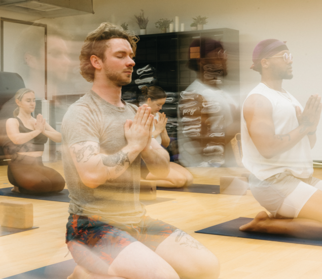 A group of people kneel on yoga mats indoors at yoga classes in Austin, eyes closed and hands in prayer position, with a blurred motion effect suggesting calm and movement. Shelves with folded blankets are in the background.