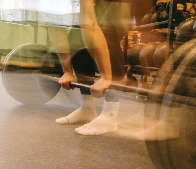 A person with blurred motion bends to grip a weighted barbell in a gym, preparing to lift. Kettlebells and gym equipment are visible in the background, highlighting the dynamic atmosphere typical of yoga classes Austin offers.
