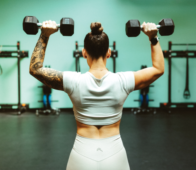A woman with a tattooed arm and hair in a bun lifts dumbbells overhead in a gym, facing away from the camera. She wears a white crop top and leggings. Fitness equipment is visible, reflecting the active vibe of yoga classes Austin offers.