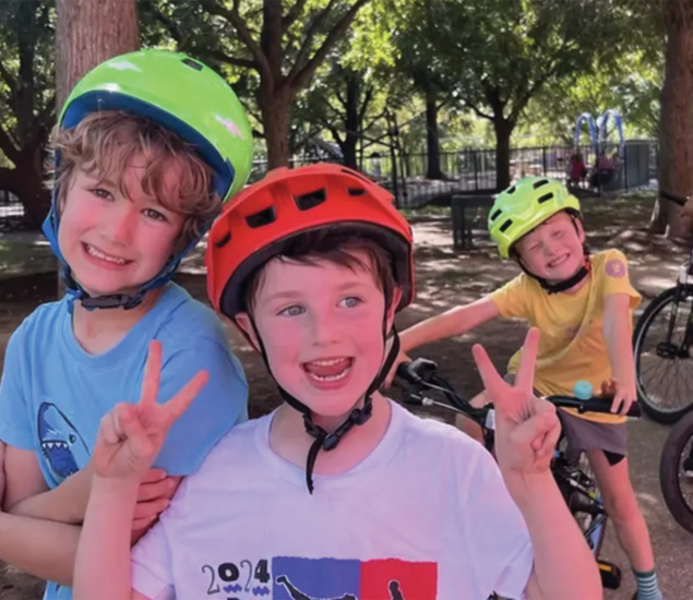 Three children wearing helmets smile at the camera and make peace signs with their hands while standing near bicycles in a park—ready for adventures like those at Austin summer camps, with trees and playground equipment in the background.