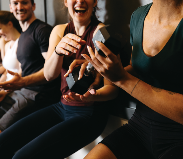 Four people in athletic wear sit together, smiling and laughing. Two of them are holding and passing a dumbbell, creating a lighthearted vibe that’s common in yoga classes Austin locals love for their friendly and energetic community.