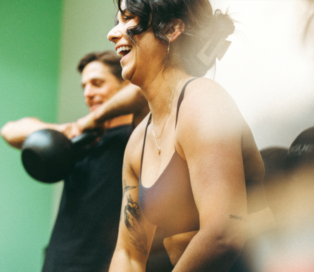 A woman smiling and laughing during a workout, with a man in the background lifting a kettlebell. Both appear to be in a gym setting, enjoying their exercise—capturing the energy often found in yoga classes Austin locals love.