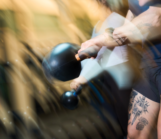 A tattooed person holds a kettlebell while exercising indoors, with motion blur emphasizing movement. Another person with a kettlebell is visible in the background, capturing the dynamic energy often found in yoga classes Austin offers.