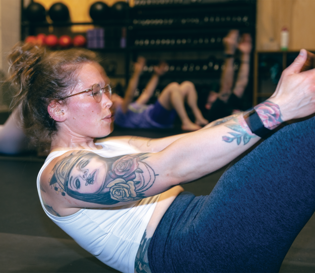 A woman with glasses and tattoos works out in a gym, performing a core exercise on a mat. In the background, others join in, reflecting the energy of yoga classes Austin is known for. Gym equipment is visible on shelves behind them.