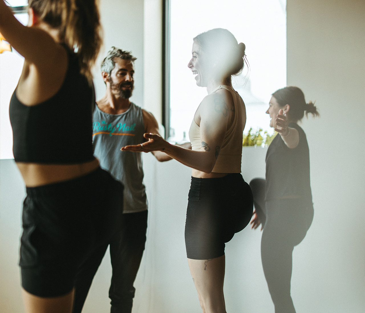 Three people in workout clothes chat by a bright window in a gym or studio, where yoga classes Austin locals love are held. One woman gestures animatedly while another enjoys a standing stretch, creating a friendly and relaxed atmosphere.