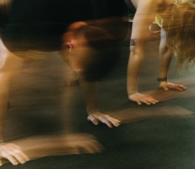 Two people doing push-ups indoors on a dark floor, with motion blur capturing their energetic movement—perfect inspiration for anyone seeking dynamic yoga classes Austin style. Visible tattoos add a unique flair to the vibrant scene.