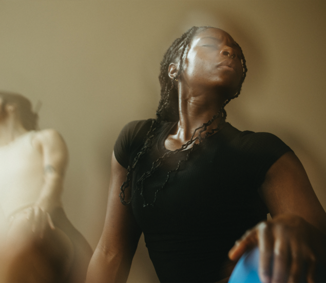 A woman with braided hair and a nose ring, wearing a black shirt, sits with her eyes closed and head tilted back, appearing relaxed and reflective—an atmosphere reminiscent of yoga classes Austin locals enjoy. Another out-of-focus person is in the background.