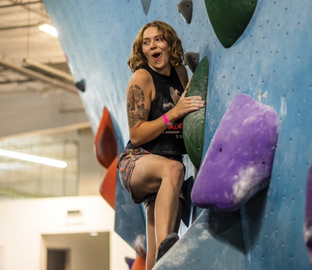 A person with short hair and tattoos climbs an indoor rock wall at a fitness classes Austin gym, smiling with excitement. They grip green and purple holds, wearing a sleeveless top and patterned shorts amid bright lights and colorful holds.