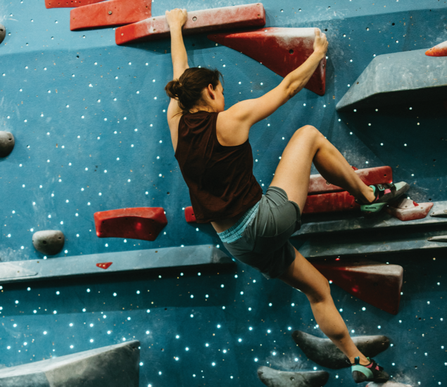 A woman wearing athletic clothing climbs an indoor bouldering wall with red and gray holds, gripping with both hands and balancing on footholds during one of the popular climbing classes in Austin.