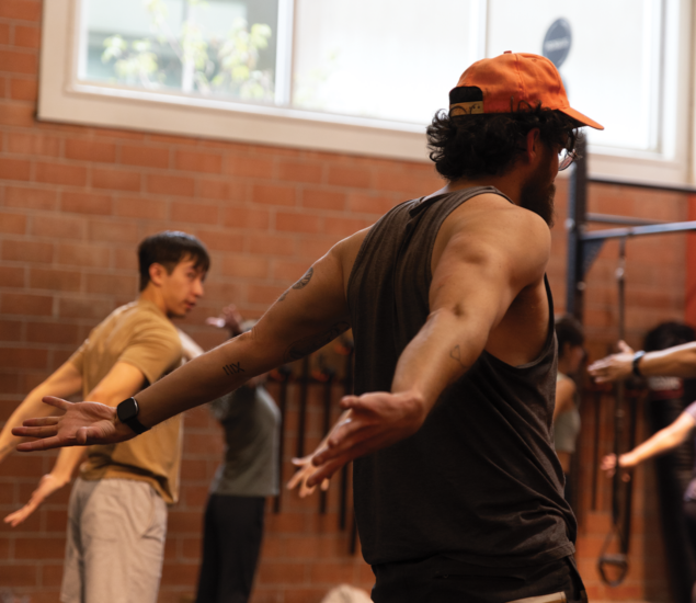 Several people stand with arms extended in a fitness studio, following an instructor in a sleeveless shirt and orange cap. The room has brick walls, workout equipment, and natural light—perfect for energizing yoga classes Austin locals love.