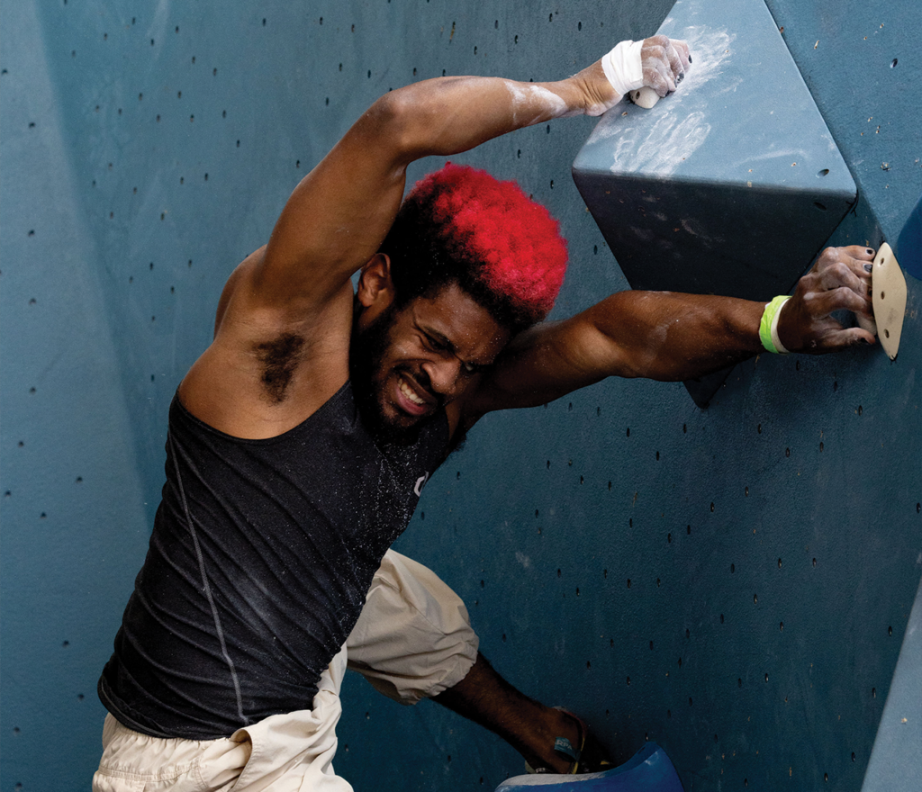 A man with red and black hair climbs an indoor bouldering wall at a fitness classes Austin gym, gripping holds with chalked hands, wearing a black tank top and light pants, with a determined expression.
