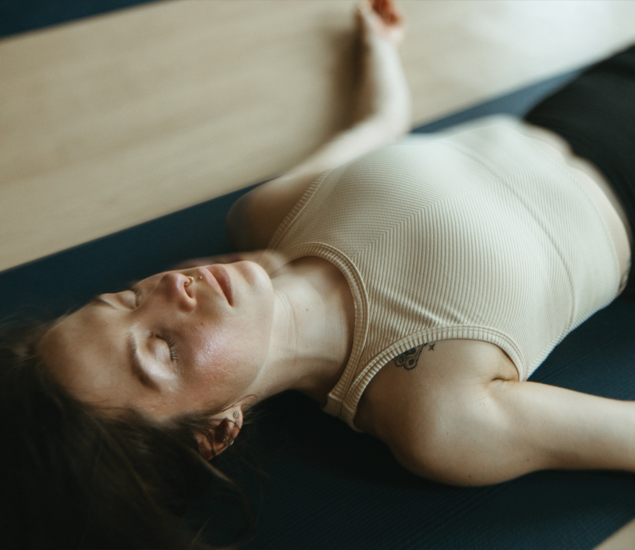 A woman in a beige tank top lies on a yoga mat with eyes closed, appearing relaxed. Her arms rest at her sides as she practices a calming pose, enjoying a peaceful moment that reflects the tranquility found in yoga classes Austin offers.