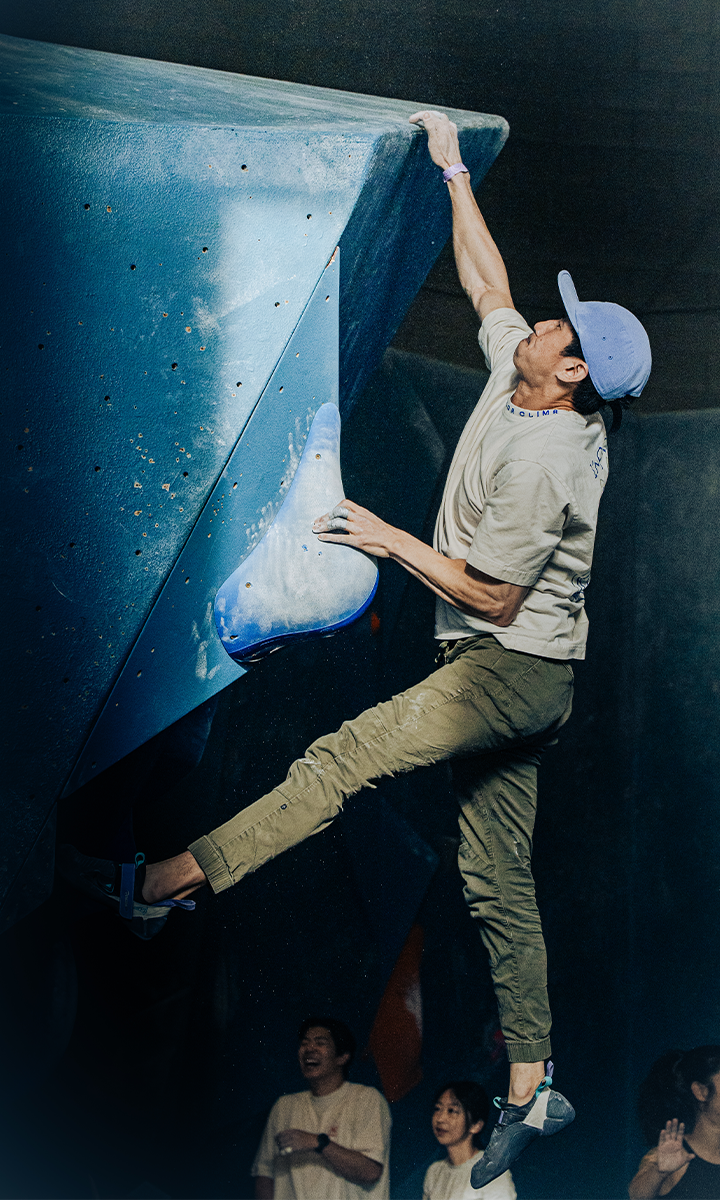 A climber wearing a cap and chalk on his hands stretches to grip the top edge of a blue wall at an indoor rock climbing gym, while spectators watch from below.