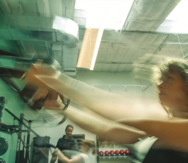 A blurred action shot of a person using suspension training straps in a gym, with another person observing in the background—capturing the intensity and movement found in dynamic workouts like yoga classes Austin offers.
