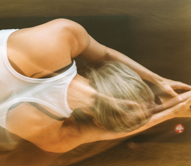 A person with blonde hair in a white tank top stretches forward on a dark surface, with a blurred double exposure effect showing movement—reflecting the energy found in yoga classes Austin offers.