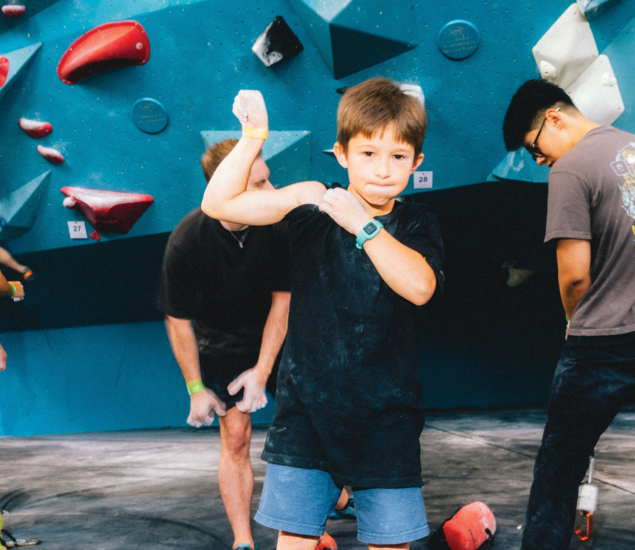 A young boy in a blue t-shirt flexes his arm and shows his muscles while standing in front of a climbing wall, capturing the fun spirit of fitness classes Austin has to offer. People nearby prepare to climb against the blue wall with red holds.