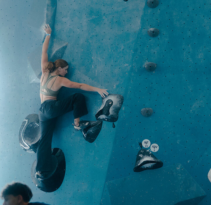 A woman climbs a blue indoor bouldering wall as four people on the ground watch her during a Seattle climbing competition. Sunlight streams through large windows in the background, creating a bright, airy atmosphere.