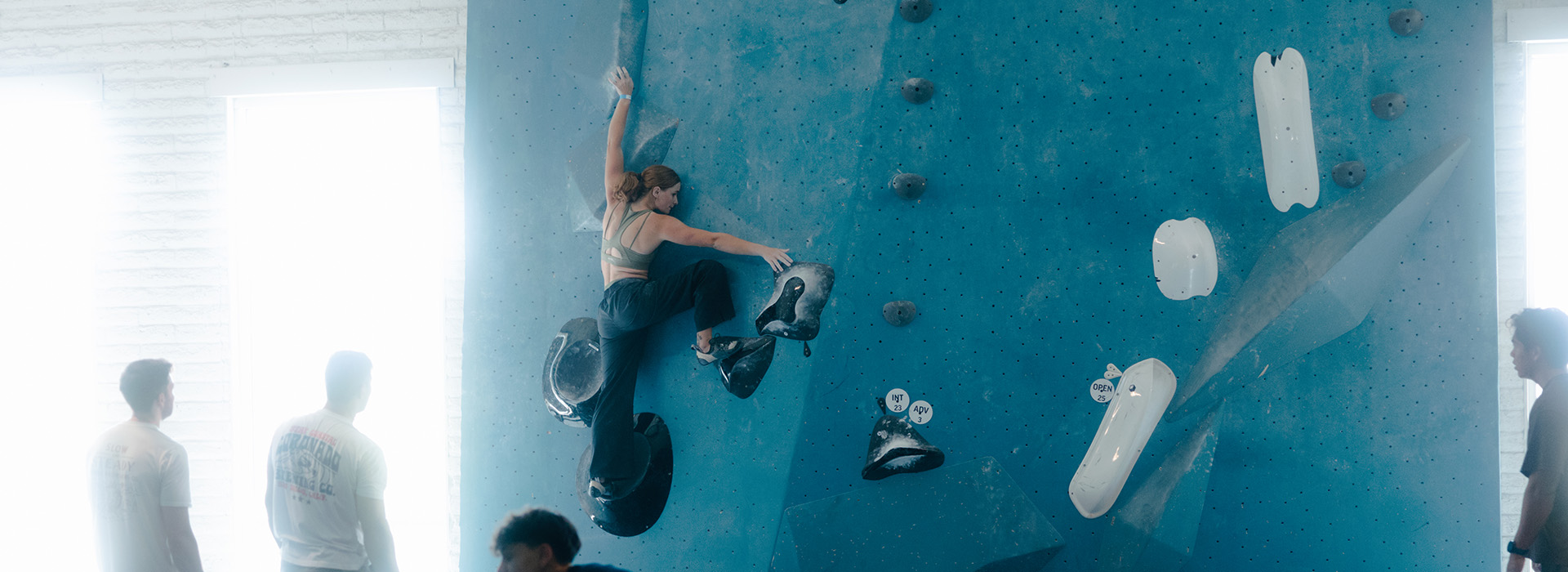 A woman climbs a blue indoor bouldering wall as four people on the ground watch her during a Seattle climbing competition. Sunlight streams through large windows in the background, creating a bright, airy atmosphere.
