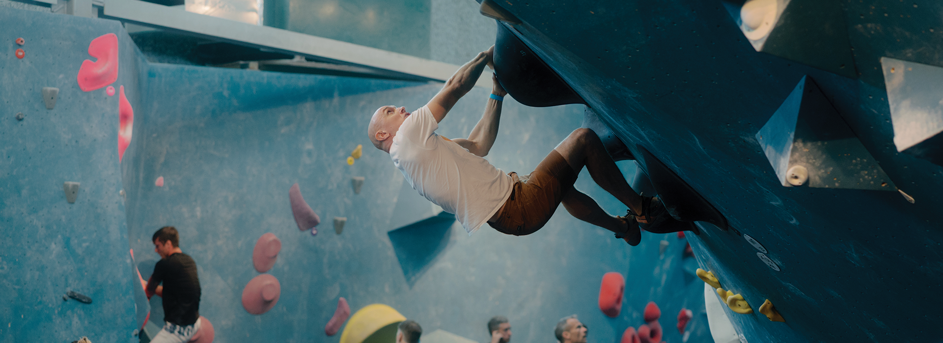A man in a white shirt and brown shorts climbs a steep overhanging wall at an indoor bouldering gym in Austin, with other climbers, colorful holds, and fitness classes Austin signs visible in the background.