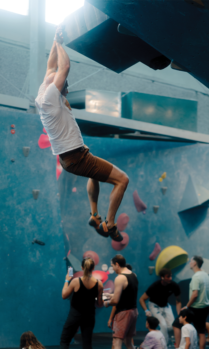 A man in a white shirt and brown shorts hangs from a climbing wall at an indoor rock climbing gym in Austin, while several people—perhaps after their fitness classes—watch and take photos below.