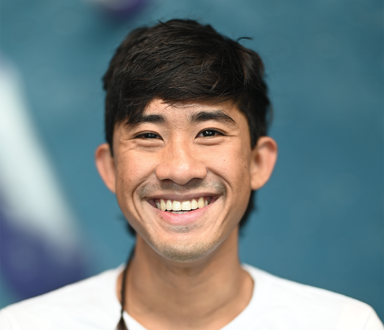 A young man with short dark hair smiles broadly at the camera, wearing a white shirt. The blurred blue and white background hints at his enthusiasm for Personal Climbing Coaching Austin.