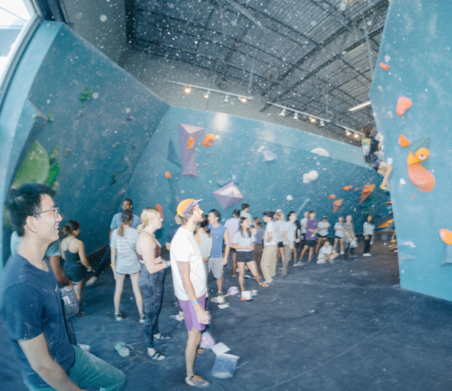 A group of people stand in line at an indoor rock climbing gym, watching climbers on blue climbing walls with colorful holds. The gym, known for its lively energy, also offers climbing classes for all skill levels.