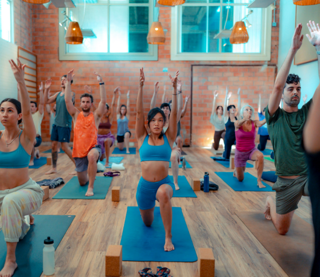 A diverse group attends Yoga Classes in a bright studio, performing a lunge pose with arms raised. They are focused and aligned on yoga mats, surrounded by wooden floors and brick walls—a welcoming space for all markets.