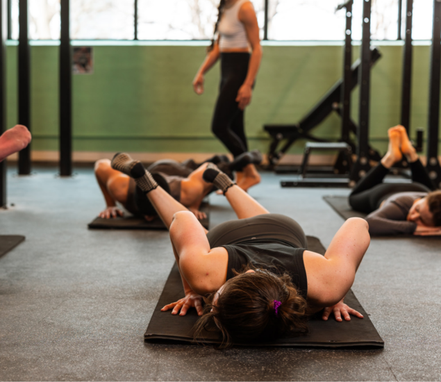 Several people are lying face down on exercise mats in a gym during fitness classes, stretching as a woman instructs the group. Gym equipment and windows can be seen in the background, highlighting an active workout scene for all markets.