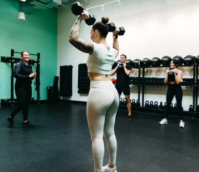 A woman in workout clothes lifts dumbbells overhead in a fitness class, while three others follow her lead. Exercise equipment and kettlebells line the walls, reflecting the variety available in all markets.