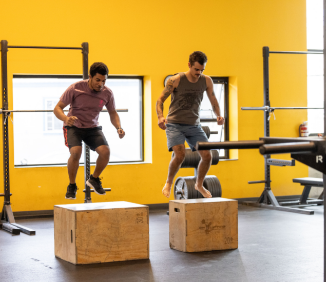 Two men are mid-air, jumping onto wooden boxes in a gym with yellow walls during a fitness class. They appear focused and athletic, wearing casual workout clothes, while barbells and racks fill the background.