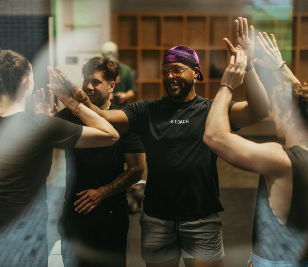 A group of people, including a smiling person wearing a COACH shirt, stand in a gym giving high-fives, celebrating as a team after an energizing fitness class. This positive energy is shared across all markets.