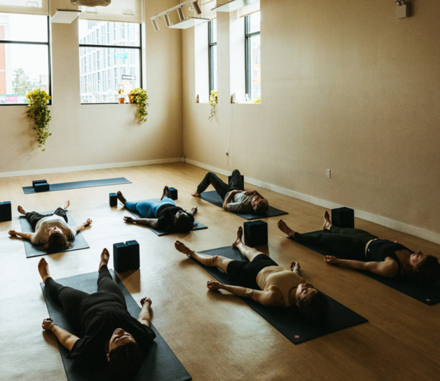 Six people lie on yoga mats in a spacious, sunlit studio during one of the Yoga Classes. Yoga blocks are placed beside them, and large windows let in natural light, highlighting the calm, peaceful atmosphere enjoyed by All Markets.