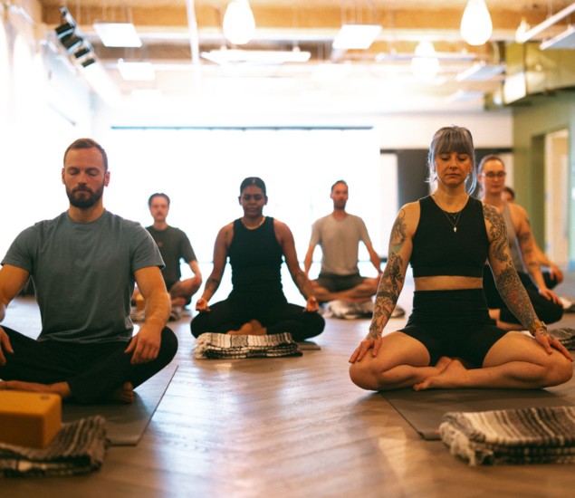 A group of people sit cross-legged on yoga mats in a bright studio, meditating during Yoga Classes with eyes closed and hands resting on their knees. The room has wooden floors, soft lighting, and blankets placed beside each person.